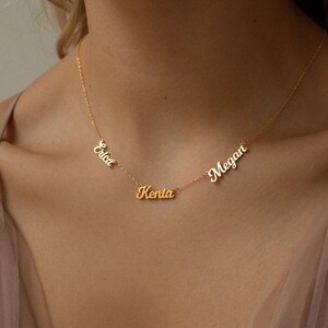 Puede incluir: Un collar dorado con tres colgantes con nombres: "Erica", "Kenia" y "Megan". Los nombres están en una fuente cursiva. El collar se lleva alrededor del cuello.