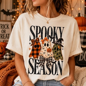 Peut inclure: Un t-shirt couleur crème avec le texte SPOOKY SEASON en lettres noires. Le motif comprend trois fantômes : un avec un motif à carreaux orange et noir, un blanc avec de petites citrouilles orange et un vert olive avec des pois noirs.