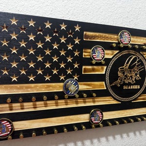 Puede incluir: Exhibidor de bandera americana de madera con estrellas y rayas talladas, con un emblema circular con la palabra "SEABEES" y varias monedas de desafío militar. La bandera tiene agujeros para exhibir artículos.