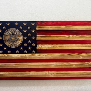 May include: Wooden American flag with the United States Army emblem. The flag features a blue canton with gold stars and the Army emblem, and red and natural wood stripes. The flag is made of wood and has a rustic appearance.