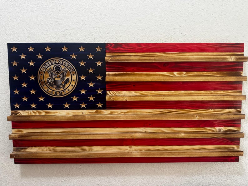 May include: Wooden American flag with the United States Army emblem. The flag features a blue canton with gold stars and the Army emblem, and red and natural wood stripes. The flag is made of wood and has a rustic appearance.