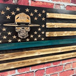 May include: Wooden American flag with a carved skull wearing a U.S. Border Patrol hat. The flag features a black background with carved stars, natural wood stripes, and a teal stripe. A unique patriotic wall decor.
