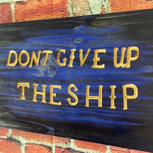 May include: A wooden sign with a blue and black background and gold lettering. The sign reads "Don't Give Up The Ship".
