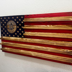 May include: Wooden American flag with red and natural wood stripes. The upper left corner features a blue field with gold stars and a gold emblem. The flag is mounted on a wall.