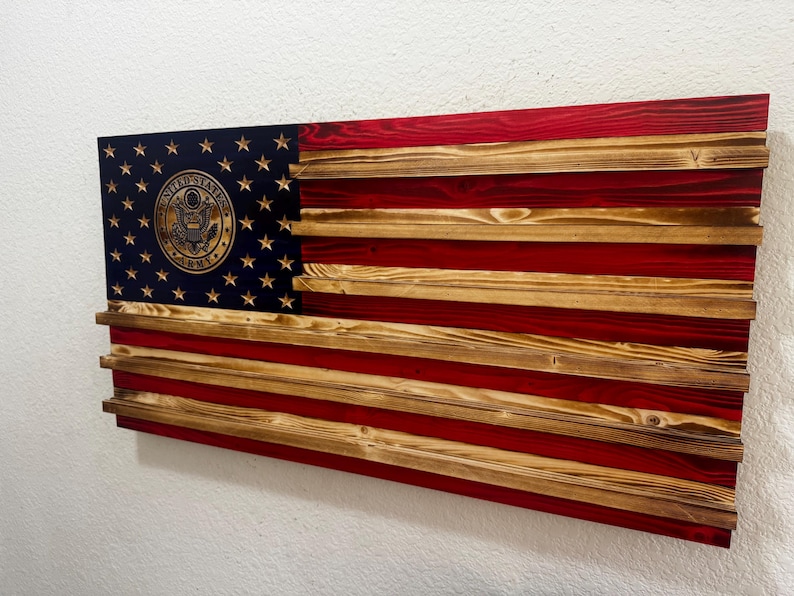 May include: Wooden American flag with red and natural wood stripes. The upper left corner features a blue field with gold stars and a gold emblem. The flag is mounted on a wall.