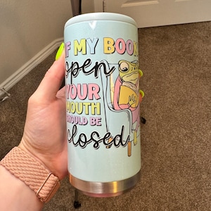 Puede incluir: Un vaso de acero inoxidable azul claro con un acabado brillante. Presenta una rana de dibujos animados sentada en una silla leyendo un libro. El texto en el vaso dice "Si mi libro está abierto, tu boca debe estar cerrada."