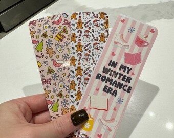 May include: Three pink and white patterned bookmarks with a winter theme. One bookmark has a gingerbread man and snowflake pattern, another has a snowman and candy cane pattern, and the third has a pink and white striped pattern with the text "In My Winter Romance Era" and a snowflake, ice skate, and book illustration.