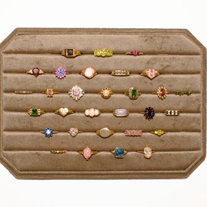 Puede incluir: Un expositor de anillos de terciopelo marrón con 25 anillos de oro con piedras preciosas de varios colores. Los anillos están dispuestos en filas en el expositor.