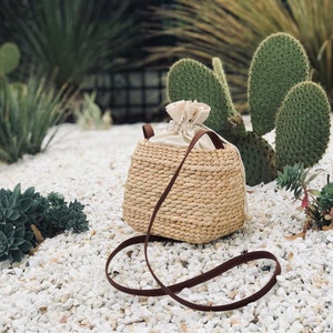 Puede incluir: Un bolso de paja tejido con una correa de cuero marr&oacute;n y un forro de algod&oacute;n blanco. El bolso est&aacute; sentado en una cama de grava blanca.