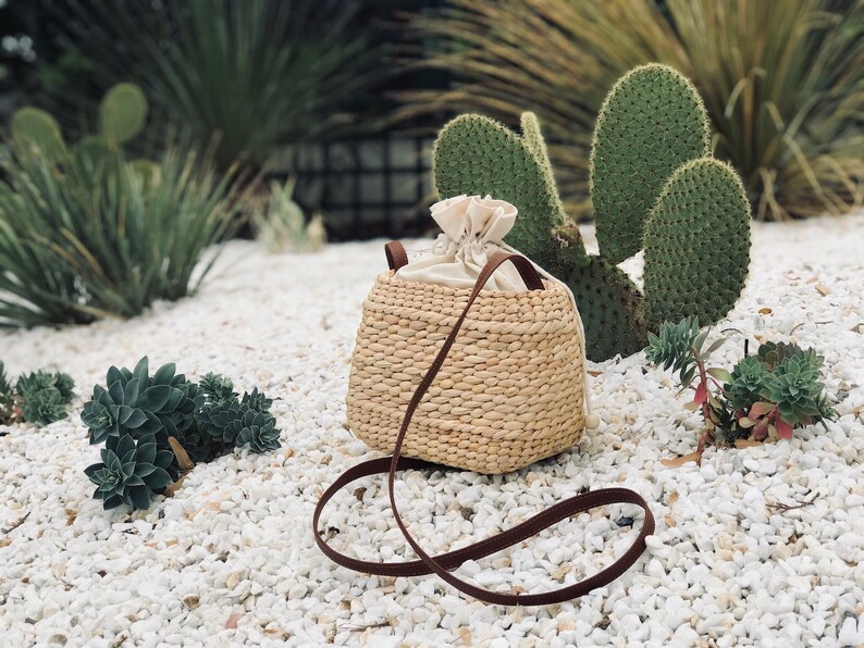 Puede incluir: Un bolso de paja tejido con una correa de cuero marr&oacute;n y un forro de algod&oacute;n blanco. El bolso est&aacute; sentado en una cama de grava blanca.