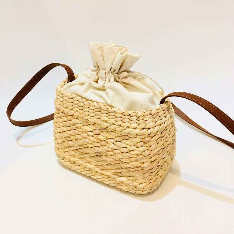 Puede incluir: Un peque&ntilde;o bolso de paja con una correa de cuero marr&oacute;n y un forro de algod&oacute;n blanco. El bolso tiene un cierre de cord&oacute;n.
