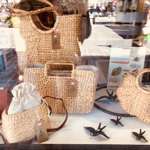 Puede incluir: Cuatro bolsas de mimbre con asas de cuero marr&oacute;n y una etiqueta de papel marr&oacute;n con el logotipo "AMBEET" en cada bolsa. Las bolsas se exhiben en un escaparate con un cartel en el fondo que dice "AMBEET" y tiene una imagen de un cangrejo.