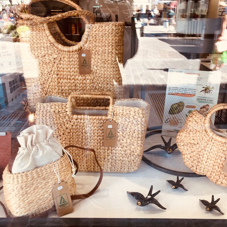 Puede incluir: Cuatro bolsas de mimbre con asas de cuero marr&oacute;n y una etiqueta de papel marr&oacute;n con el logotipo "AMBEET" en cada bolsa. Las bolsas se exhiben en un escaparate con un cartel en el fondo que dice "AMBEET" y tiene una imagen de un cangrejo.