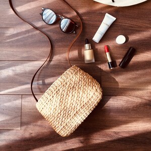 Puede incluir: Un bolso de paja con una correa de cuero marr&oacute;n, gafas de sol, una botella de suero, un tubo de crema facial La Mer, un peque&ntilde;o recipiente redondo y un l&aacute;piz labial rojo sobre una superficie de madera.