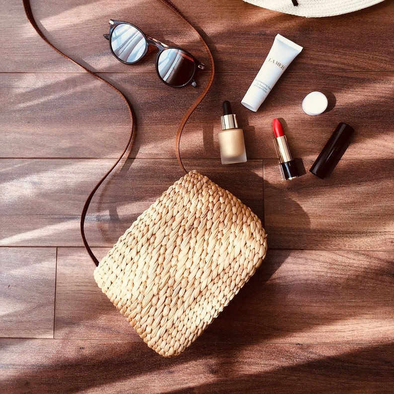 Puede incluir: Un bolso de paja con una correa de cuero marr&oacute;n, gafas de sol, una botella de suero, un tubo de crema facial La Mer, un peque&ntilde;o recipiente redondo y un l&aacute;piz labial rojo sobre una superficie de madera.