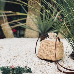 Puede incluir: Un bolso de paja tejido de color marr&oacute;n claro con una correa de cuero marr&oacute;n y un forro de tela blanco. El bolso est&aacute; sentado en una cama de guijarros blancos.