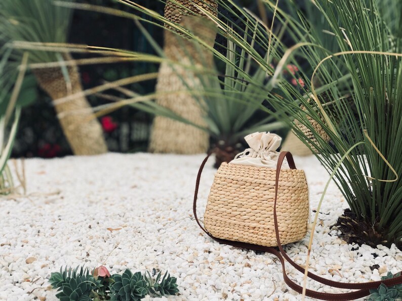 Puede incluir: Un bolso de paja tejido de color marr&oacute;n claro con una correa de cuero marr&oacute;n y un forro de tela blanco. El bolso est&aacute; sentado en una cama de guijarros blancos.