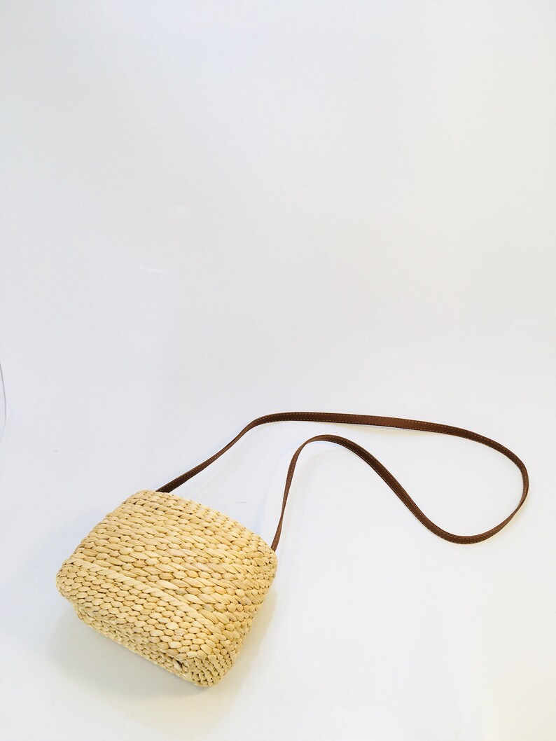 Puede incluir: Un peque&ntilde;o bolso rectangular de paja con una correa de cuero marr&oacute;n. El bolso est&aacute; tejido con paja natural y tiene un dise&ntilde;o simple y cl&aacute;sico.