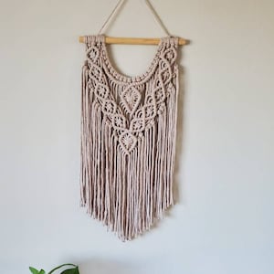 May include: A beige macrame wall hanging with a wooden dowel and hanging cord. The intricate design features knotted patterns and long fringe. A small potted plant in a blue and white patterned pot sits on a table below the wall hanging.