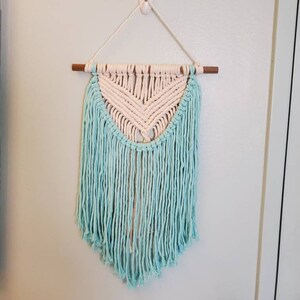 May include: A macrame wall hanging with a cream-colored chevron design and a fringe of light blue yarn. The wall decor is suspended from a wooden dowel and hangs from a white hook on a light blue wall.