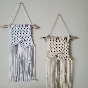 May include: Two macrame wall hangings. One is white, the other is cream-colored. Both are suspended from a wooden dowel with a triangular rope hanger. The macrame features a diamond pattern and fringe with decorative knots.