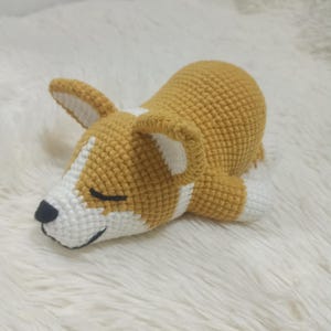 以下が含まれることがあります： 寝ているポーズの、かぎ針編みで作ったぬいぐるみ犬。犬は茶色と白で、黒い鼻と目を閉じている。