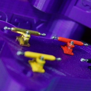 Op de afbeelding: Paarse plastic fingerboard display met het woord "CPEX" erop gedrukt. Zes verschillende gekleurde fingerboard trucks worden op het oppervlak getoond.