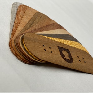 Puede incluir: Una colección de fingerboards de madera en varios patrones de veta de madera, incluyendo arce, nogal y cerezo. Los fingerboards están dispuestos en forma de abanico, con el fingerboard superior con un pequeño logotipo de un árbol estilizado.