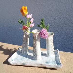 May include: A handmade ceramic flower vase with three cylindrical openings, each holding fresh flowers. The vase is white with a light blue base and decorative patterns. The flowers include an orange poppy, pink blooms, and green foliage.