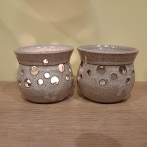 May include: Two speckled grey ceramic candle holders. Each has a rounded shape, a wide rim, and circular cutouts. The cutouts allow light from the candles inside to shine through, creating a warm glow. The candle holders are approximately 10 cm in height.