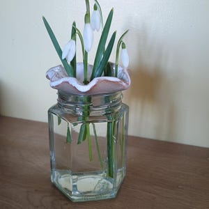 May include: A glass vase with a white ceramic top, holding a bouquet of white snowdrop flowers with green stems.