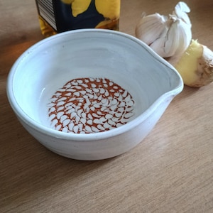 May include: A white ceramic bowl with a spout, featuring a spiral pattern in the centre. The bowl is next to a bottle of oil and a head of garlic. The bowl is likely used for food preparation.