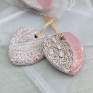 May include: Two heart-shaped ceramic ornaments with a textured, off-white lace design and a pink glazed finish. Each ornament has a hole for hanging and is tied with raffia. One is partially visible in a sheer bag.