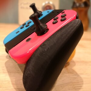May include: A pink and blue Nintendo Switch console with black joy-con controllers and black thumbstick grips. The console is attached to a black 3D printed grip.