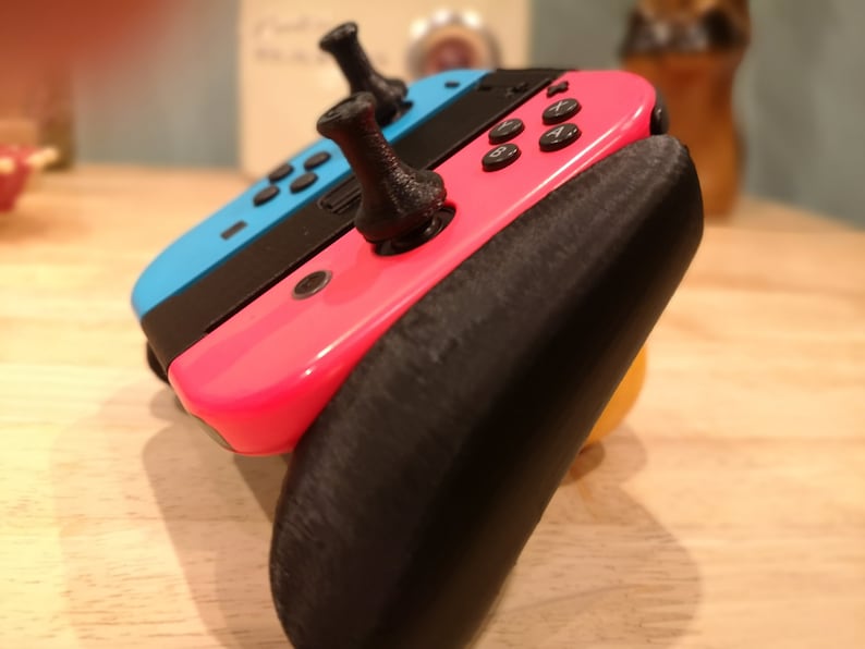 May include: A pink and blue Nintendo Switch console with black joy-con controllers and black thumbstick grips. The console is attached to a black 3D printed grip.