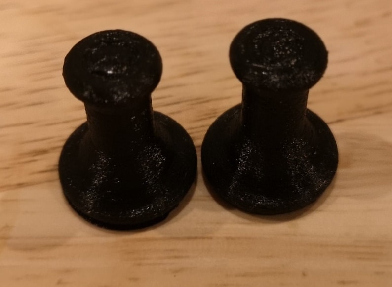 May include: Two black plastic knobs with a round top and a cylindrical base. The knobs are sitting on a wooden surface.
