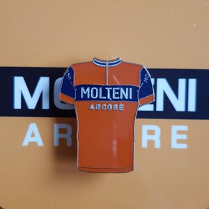 May include: Orange and blue enamel pin shaped like a cycling jersey with the text "MOLTENI ARCORE" on the front.