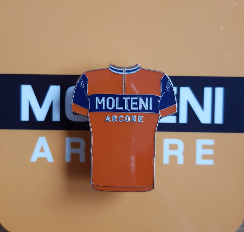 May include: Orange and blue enamel pin shaped like a cycling jersey with the text "MOLTENI ARCORE" on the front.