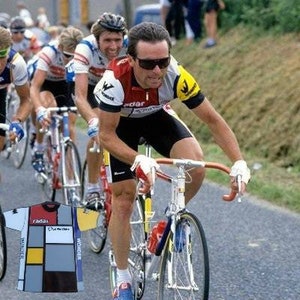 May include: A group of cyclists wearing brightly colored cycling jerseys race on a paved road. The cyclist in the foreground is wearing a red, white, and blue jersey with the word "Wonder" on the back. He is wearing sunglasses and has a determined expression on his face.