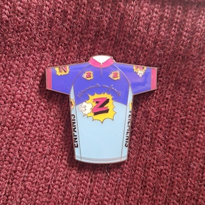 May include: A blue and pink enamel pin shaped like a cycling jersey with a cartoon 'Z' logo and the text 'V&ecirc;tements enfants' on the front. The text 'Enfants' and 'V&ecirc;tements' are on the sleeves.