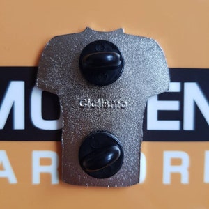 May include: A silver metal pin in the shape of a t-shirt with two black metal clasps on the back. The pin has the text "Click here" on the back.