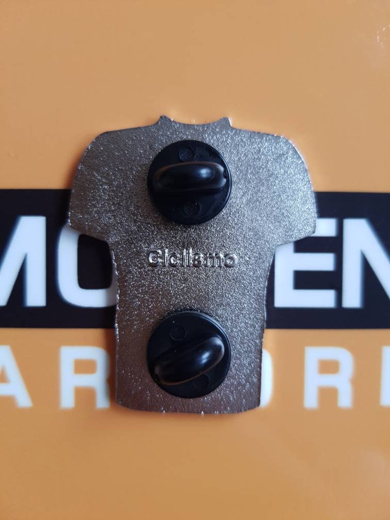 May include: A silver metal pin in the shape of a t-shirt with two black metal clasps on the back. The pin has the text "Click here" on the back.