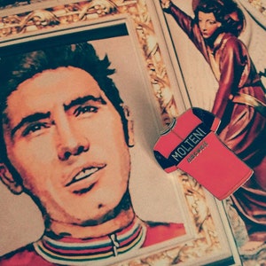 May include: A red and black cycling jersey with the text "MOLTENI" and "ARCORE" on the front. The jersey is pinned to a vintage magazine cover featuring a portrait of a man.