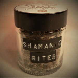 May include: A clear glass jar with a black lid, filled with dried herbs. The label reads "SHAMANIC RITES" in white letters on a black background. The jar's lid has a circular label with the text "The Forest Cauldron".