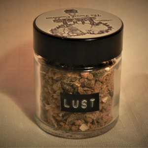 Könnte beinhalten: Ein Glasgefäß gefüllt mit braunen und grünen Kräutern, beschriftet mit "LUST". Das Gefäß hat einen schwarzen Deckel mit einem weißen Etikett, auf dem "The Forest Collection" steht.