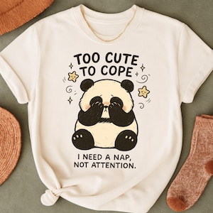 Może przedstawiać: Kremowa koszulka z ilustracją pandy zasłaniającej oczy. Tekst "TOO CUTE TO COPE" i "I NEED A NAP, NOT ATTENTION" jest wydrukowany nad i pod pandą. Projekt zawiera gwiazdy i wiry.