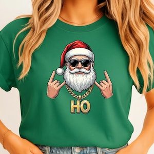 Puede incluir: Camiseta verde con un gráfico de Papá Noel con gafas de sol y una cadena de oro, haciendo un gesto de rock and roll con la mano. La palabra "HO" se muestra en letras doradas debajo.