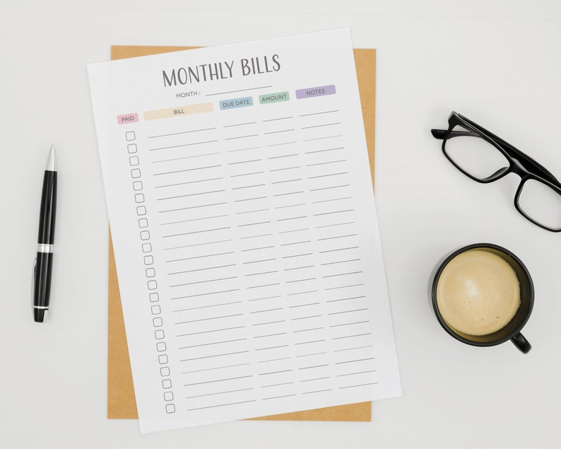 May include: A printable monthly bill tracker with columns for month, bill, due date, amount, and notes. The tracker is printed on white paper and has a brown envelope behind it. There is a black pen, a cup of coffee, and a pair of glasses on a white surface.