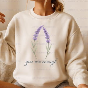 May include: Cream-colored sweatshirt featuring a lavender floral design and the phrase "you are enough" in script. The lavender illustration is in shades of purple and green. The sweatshirt has a crew neck and long sleeves.
