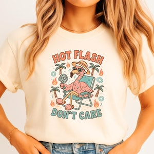 Op de afbeelding: Crèmekleurig T-shirt met een cartoon flamingo met een zonnebril en een hoed, zittend in een strandstoel. De tekst "HOT FLASH DON'T CARE" staat boven en onder de flamingo-illustratie, met palmbomen en vlammen.